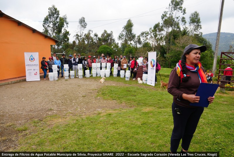 A group of community members stand together by the water filter buckets and banners showing the Water Hub and SHARE logos